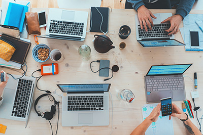 laptops, coffee cups, cords, hands all shown from above a busy work table.